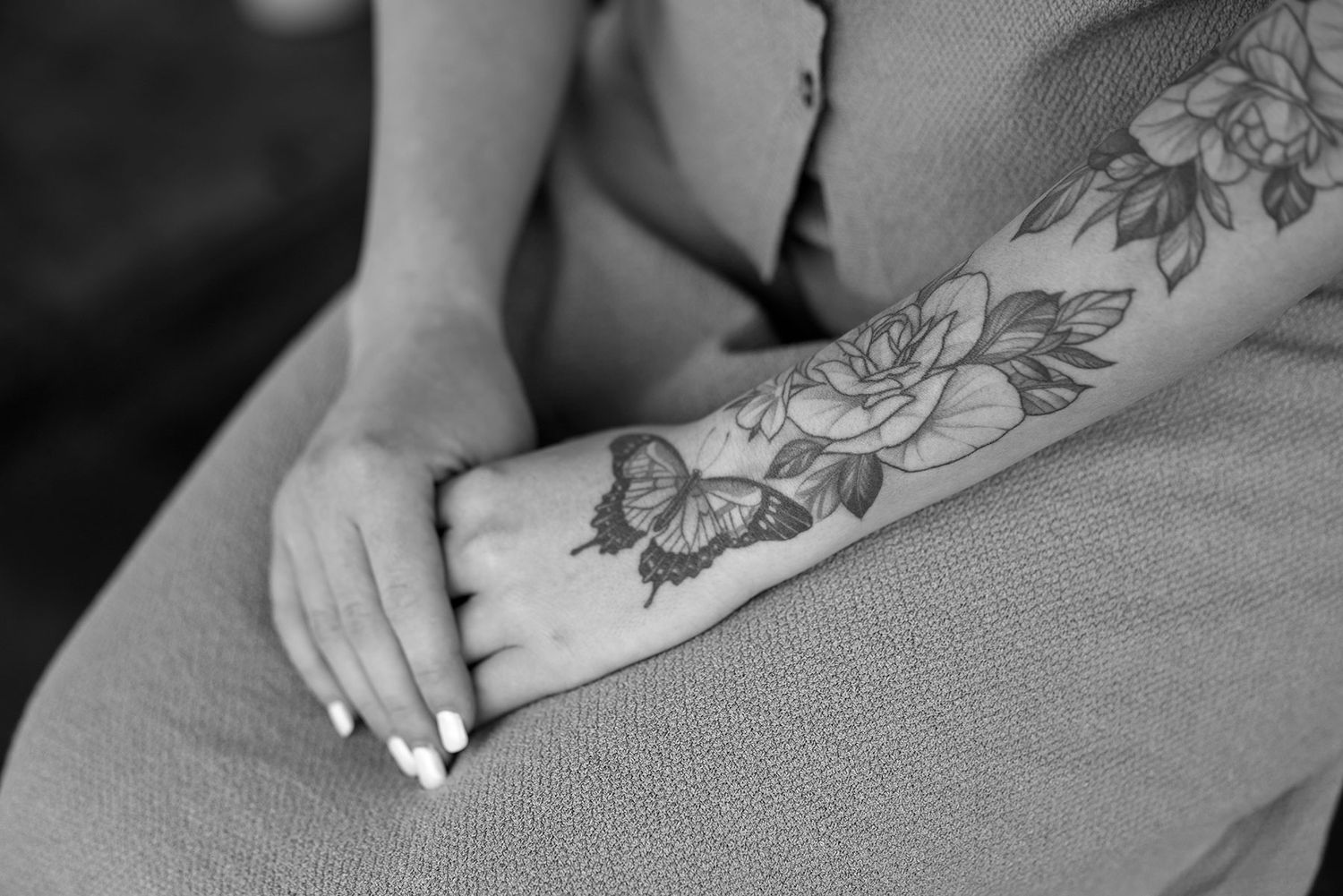 A floral tattoo design featuring delicate blooms decorates an arm resting on brown fabric with white manicured nails.