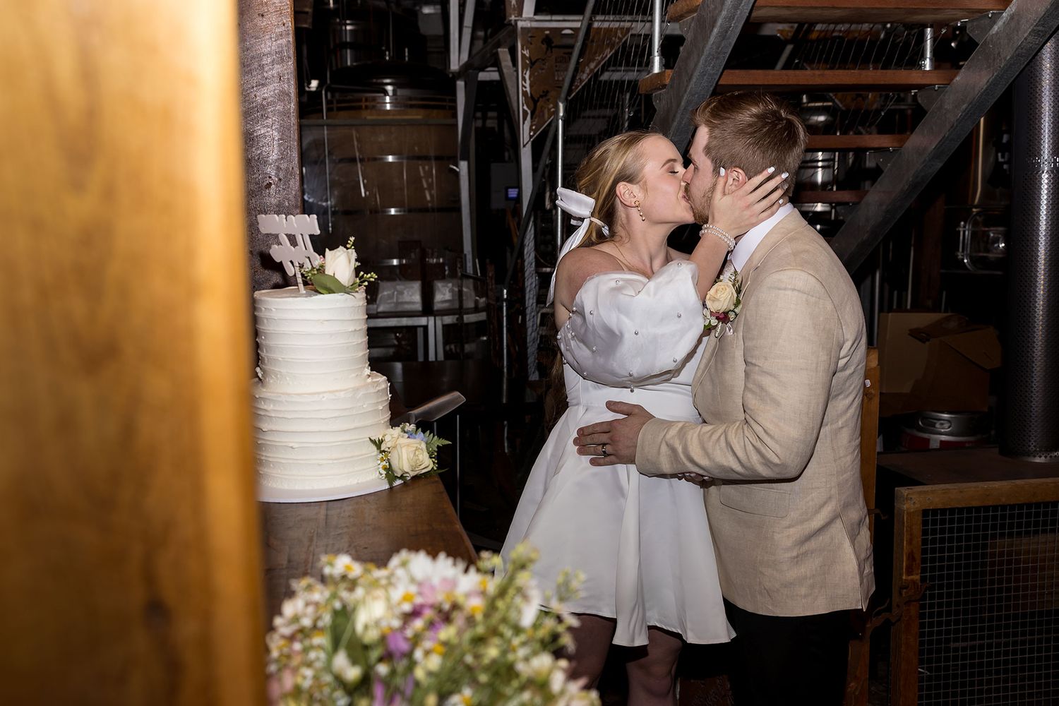 A romantic wedding moment captured beside an elegant white tiered cake with floral decorations in the rustic Bridge Road Brewers.
