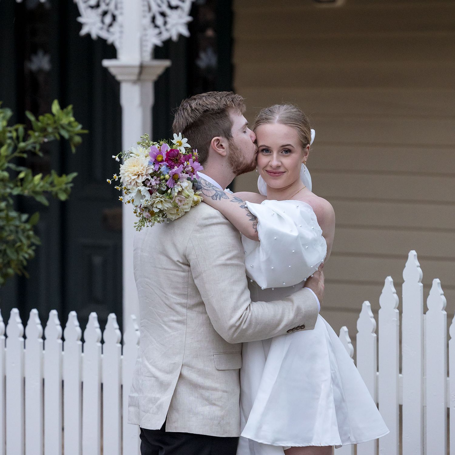 A romantic wedding moment between newlyweds in white attire standing by a picket fence and garden.