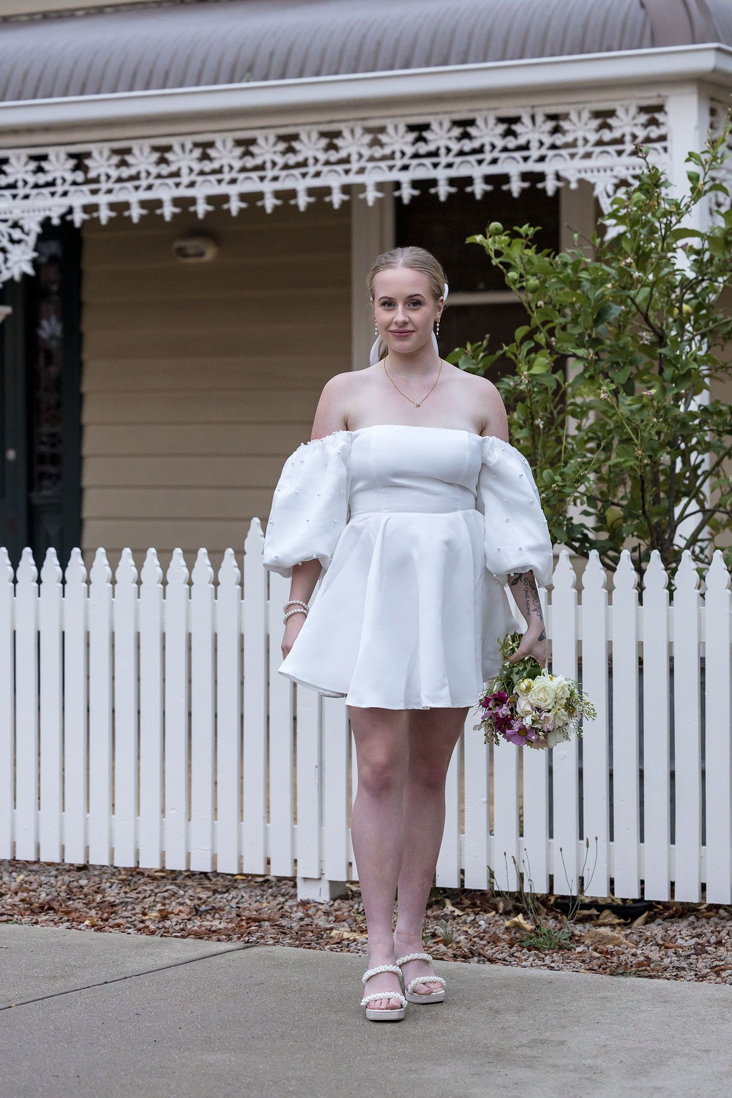 A Bride in a white off-shoulder mini dress with puffy sleeves against a picket fence and Victorian-style house veranda in Beechworth.