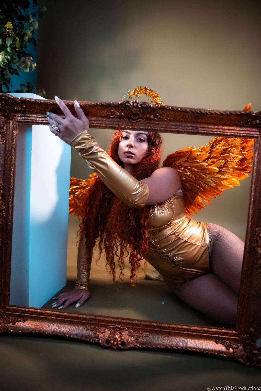 Artistic portrait in gold tones featuring large decorative wings against an ornate frame with dramatic lighting.