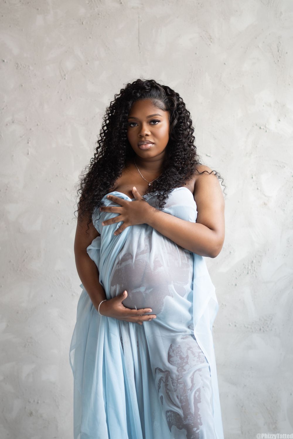 A maternity portrait in soft blue fabric against a textured gray wall.