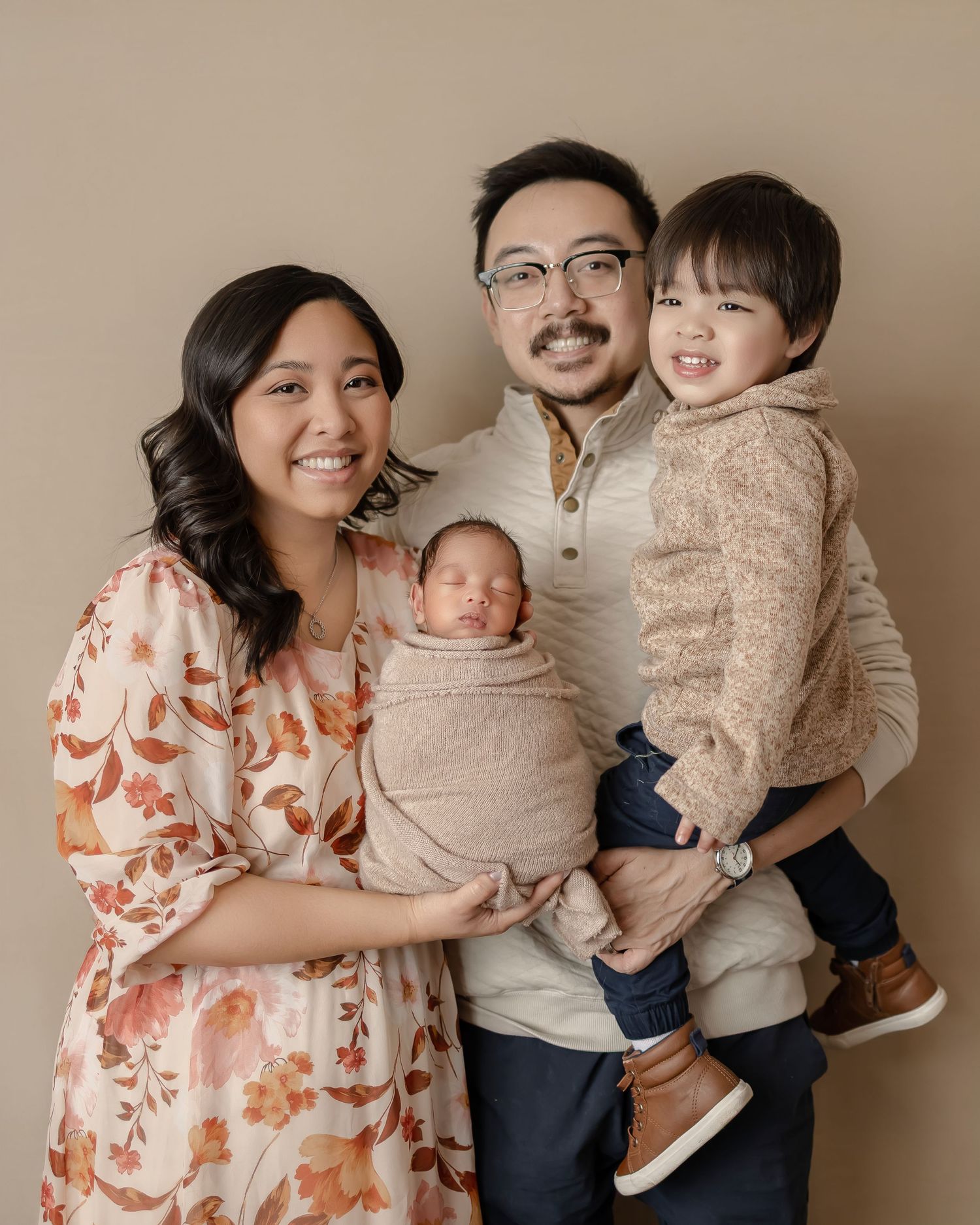 A family portrait in neutral tones featuring parents with their two children against a beige background.