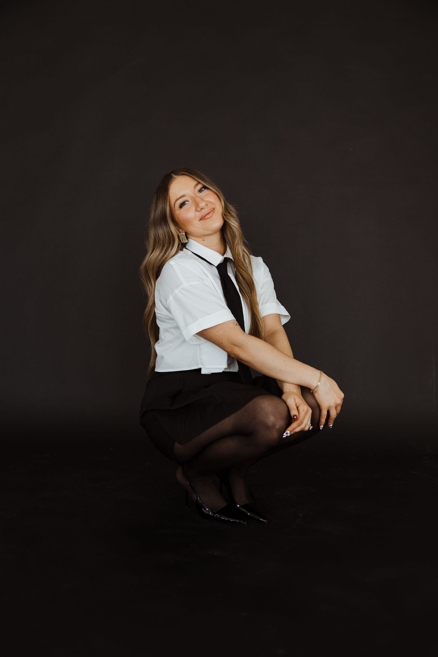 Smiling blonde woman in white shirt, black tie and skirt crouching on dark studio floor.