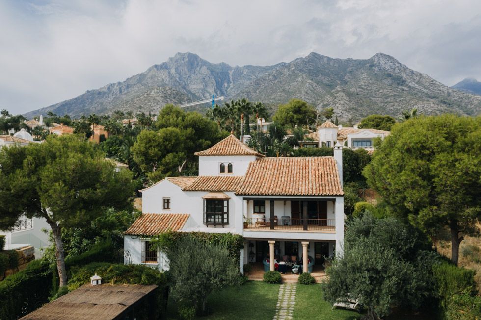 Mediterranean villa with terracotta roof nestled in lush greenery against dramatic mountain backdrop in sunny Spain.