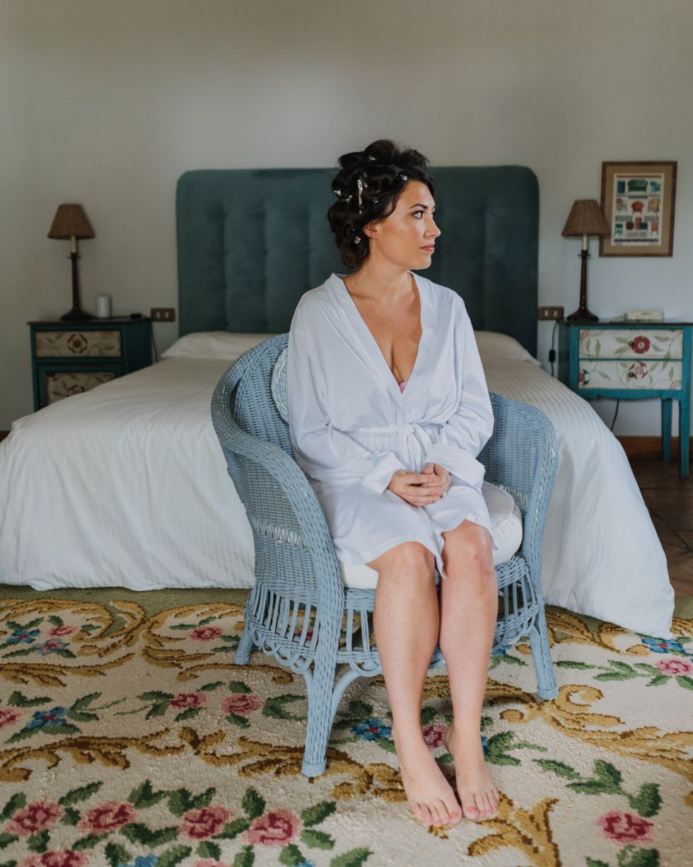 A person in a white robe sits in a blue wicker chair beside a bed on a floral rug during wedding preparations.