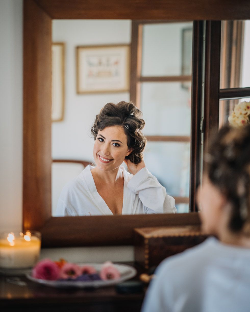 Someone sitting at a vintage vanity mirror with lit candles and flowers during wedding day preparations.