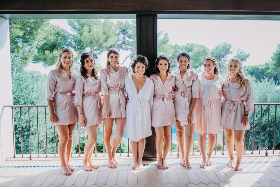 A group in matching pink robes and one in white stand together on a covered balcony with greenery in the background.