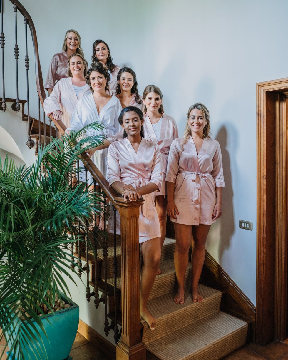 Wedding party wearing matching pink satin robes poses together on wooden staircase with potted plant nearby.