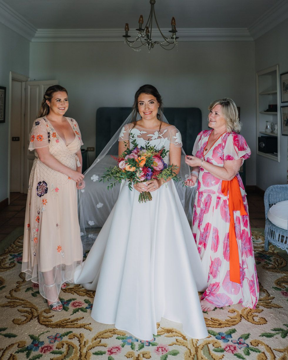 A bride in a white wedding gown poses with bridesmaids wearing pink and coral floral dresses in an elegant room with chandelier.