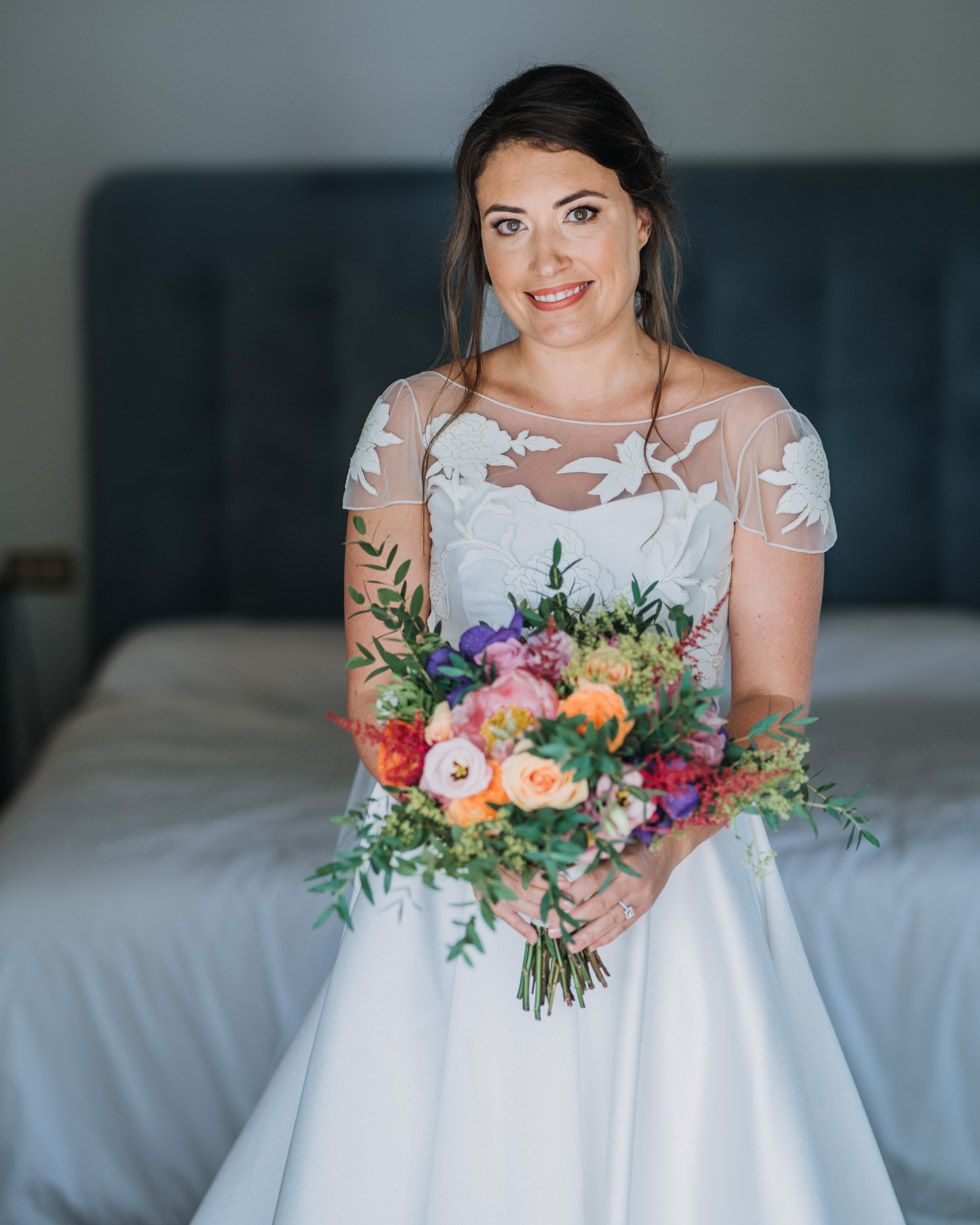 A wedding bouquet of colorful flowers including pink, orange and purple blooms held against a white gown.