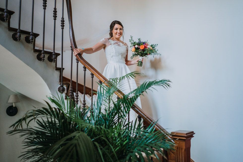 A person in a white wedding dress poses elegantly on a wooden staircase surrounded by tropical plants.