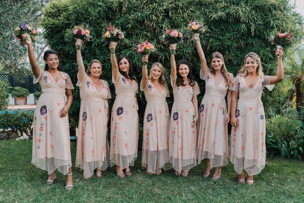 A group wearing pale pink bridesmaid dresses raise bouquets joyfully in a garden setting.