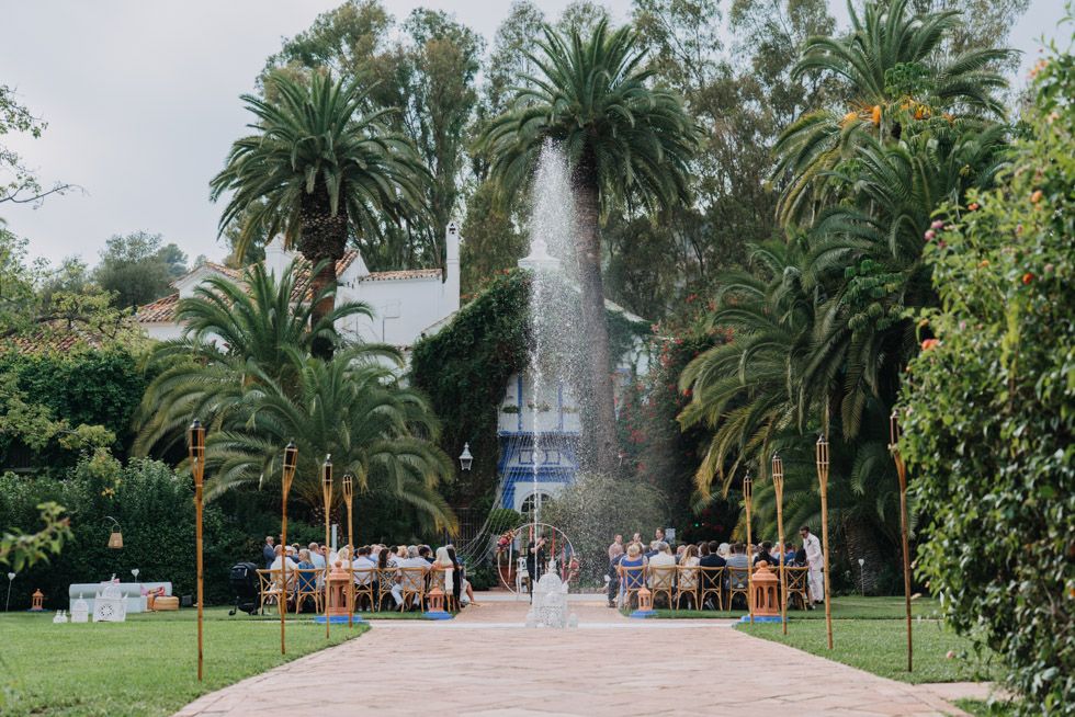 Garden pathway lined with palm trees leading to a fountain with people gathering for an outdoor event.