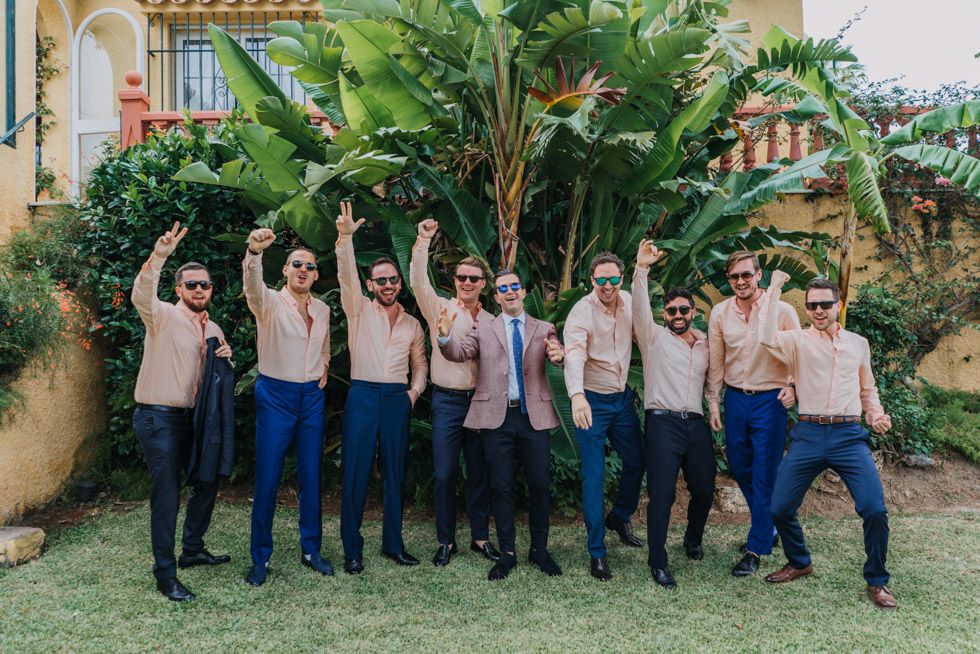 Groomsmen in navy pants and suspenders strike fun poses together in a tropical garden setting.