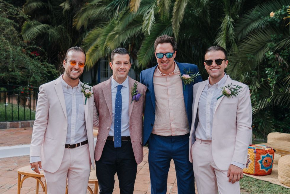 A group of four guests in formal attire pose together at an outdoor wedding with palm trees in the background.