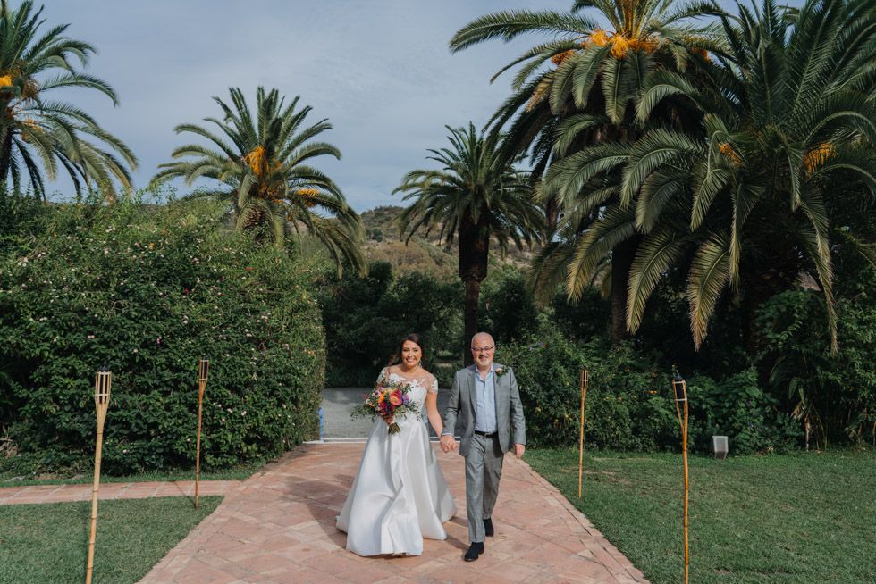 A couple walks down a palm-lined path at a tropical wedding venue on a cloudy day.