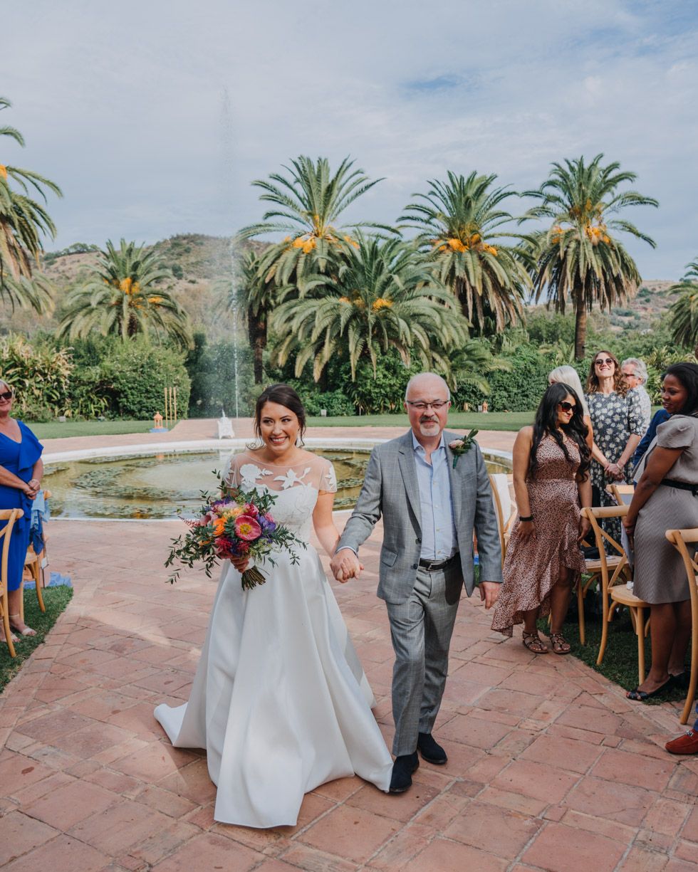 A wedding ceremony takes place outdoors with palm trees and mountains in the background on a terracotta tiled patio.