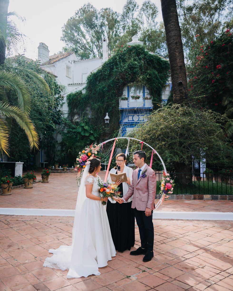 Outdoor wedding ceremony takes place at a charming venue with palm trees and lush greenery surrounding a white building.