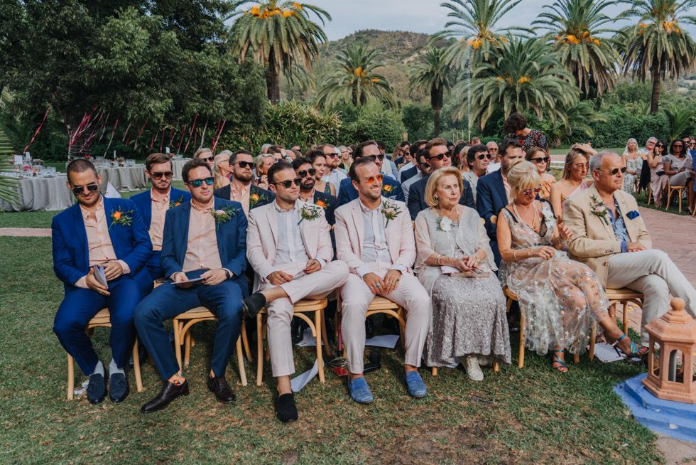 Wedding guests in coordinated neutral and blue attire seated in rows on a lawn with palm trees in the background.