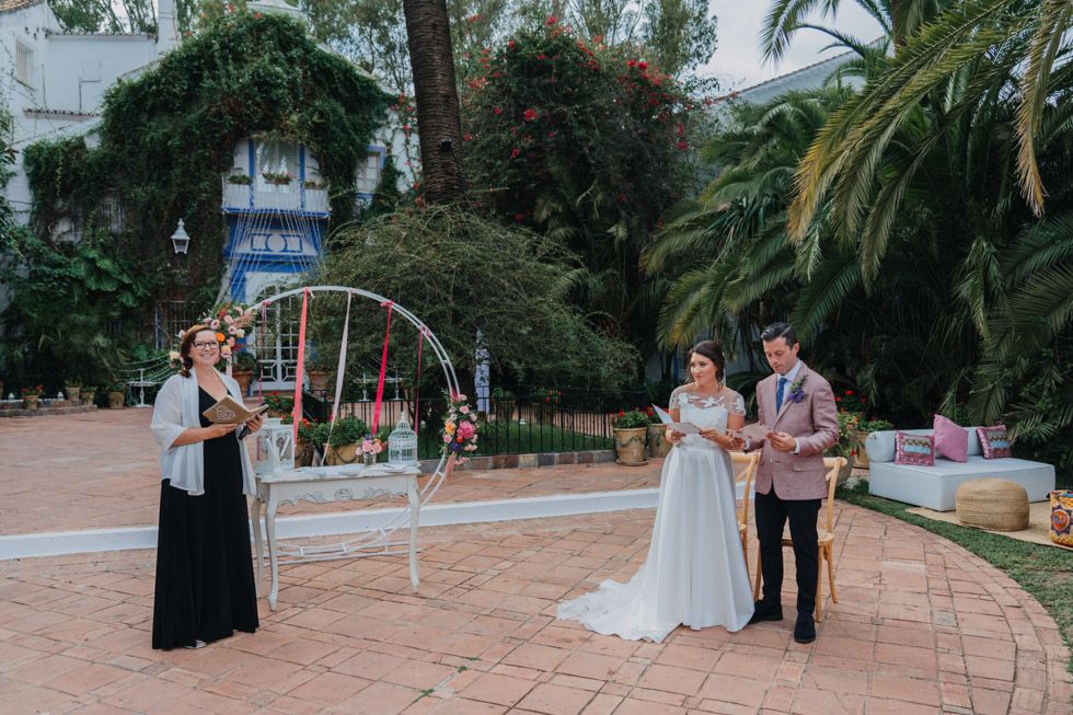 A wedding celebration takes place in a garden setting with palm trees and an arched white floral backdrop on a brick walkway.