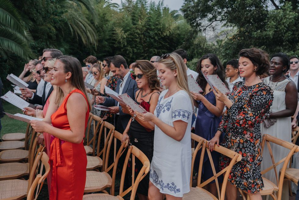 A group stands in a row at an outdoor event, looking down at papers while singing or reading together in a garden setting.