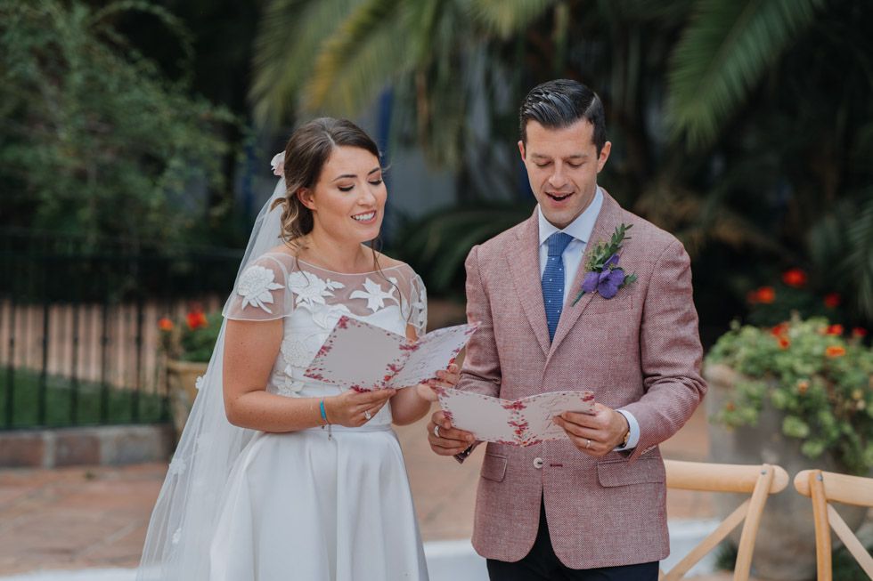 A bride and groom stand together in a garden setting reading from papers during their wedding ceremony.