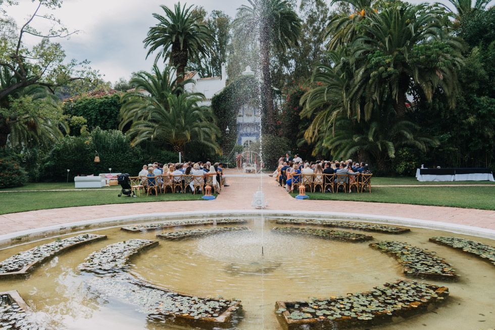 People gather around a circular fountain surrounded by palm trees in a lush garden setting.