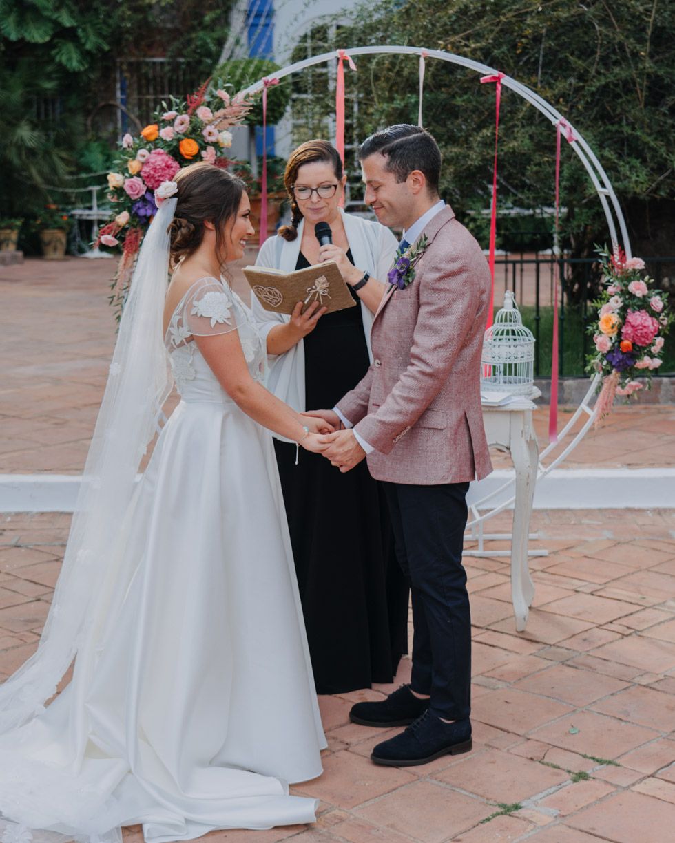 A charming outdoor wedding ceremony with a circular arch adorned with colorful flowers on a brick patio.