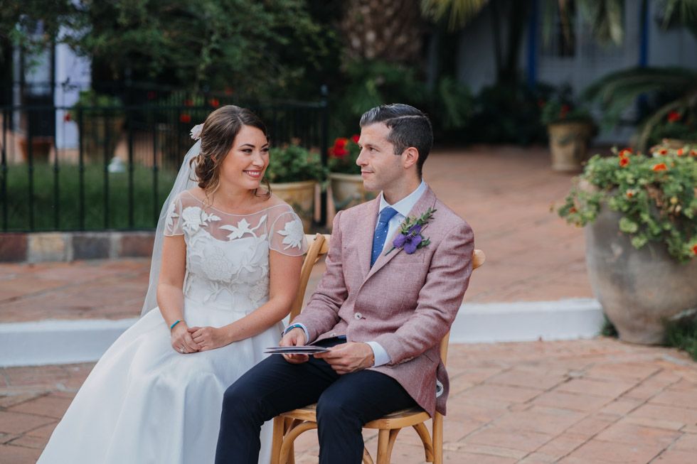 A bride and groom share a joyful moment while seated outdoors on a rustic patio surrounded by potted flowers.