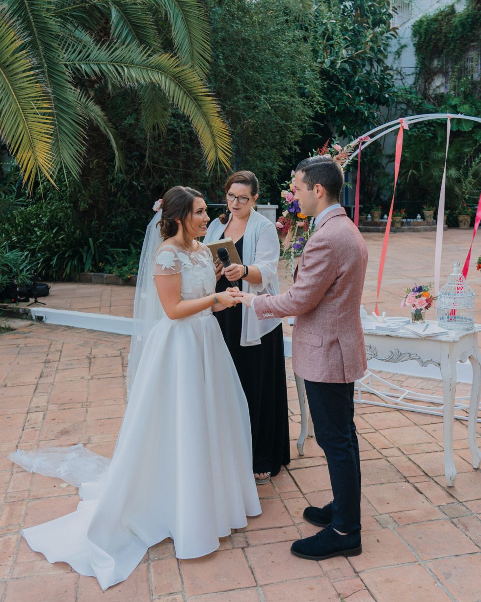 A wedding ceremony takes place outdoors under palm trees with a pink-suited groom and bride in white dress.