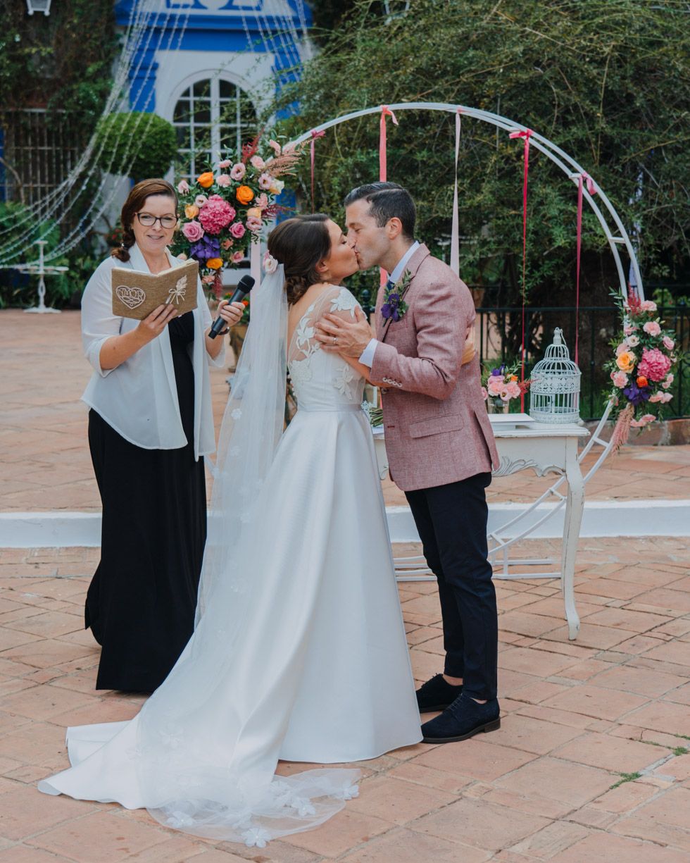 Wedding couple shares their first kiss as newlyweds under a circular floral arch with an officiant on a terrace.