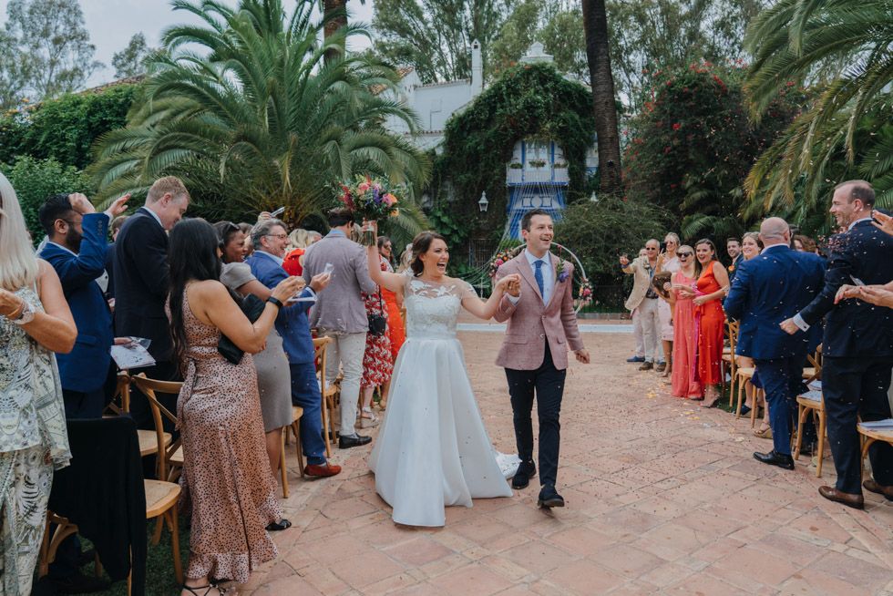 A wedding celebration in an outdoor garden setting with palm trees as guests line the pathway.