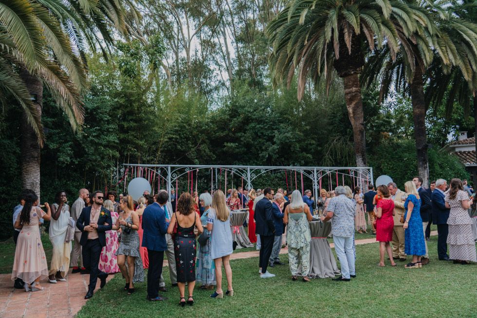 Outdoor garden wedding reception with guests gathered under palm trees and string lights on a lush green lawn.