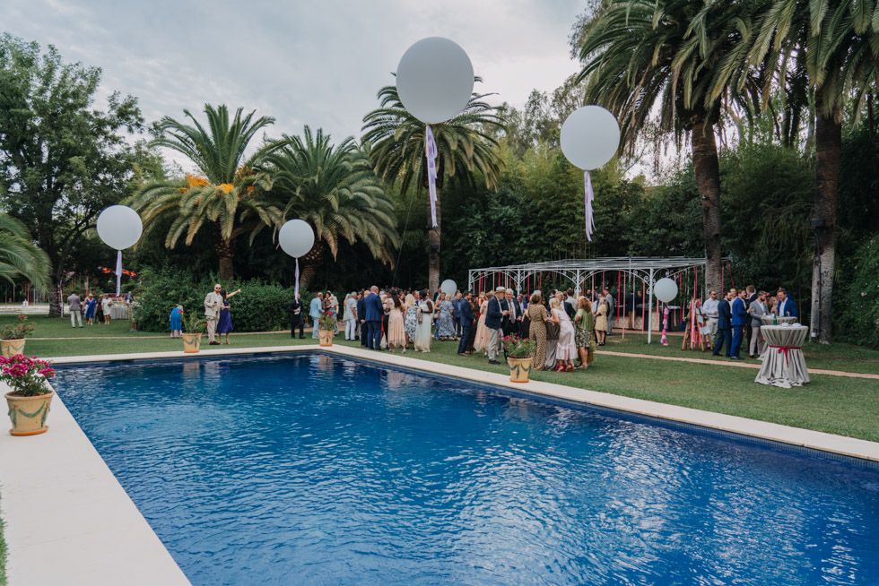 Outdoor wedding reception by a swimming pool with palm trees and white balloon decorations.