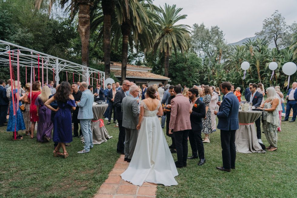 Wedding guests gather for an outdoor garden reception under palm trees and string lights.