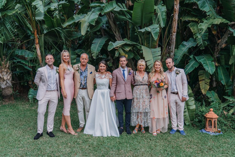 Wedding party in pastel attire poses together in front of lush tropical greenery during an outdoor garden ceremony.