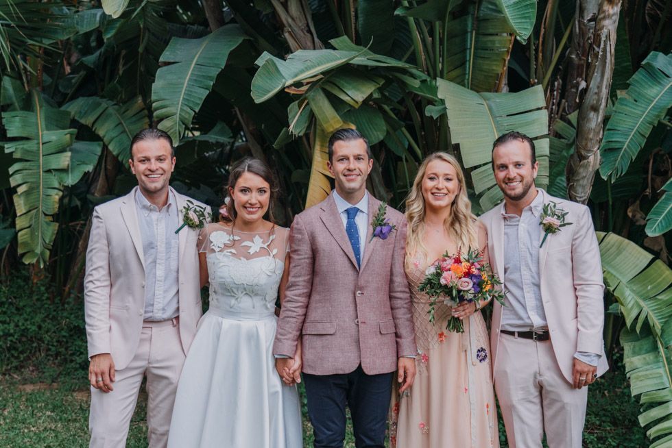 Wedding party poses outdoors in pastel attire with tropical foliage backdrop.