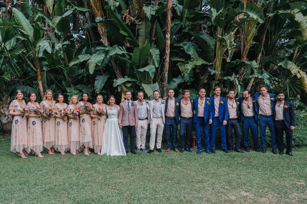 Wedding party members in blush pink and navy blue attire pose together on a lawn with tropical foliage backdrop.