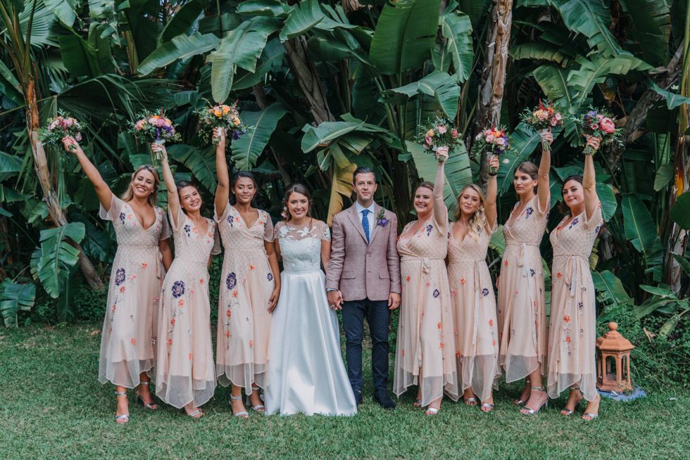 Wedding party poses in front of lush tropical foliage with bridesmaids in blush pink dresses.