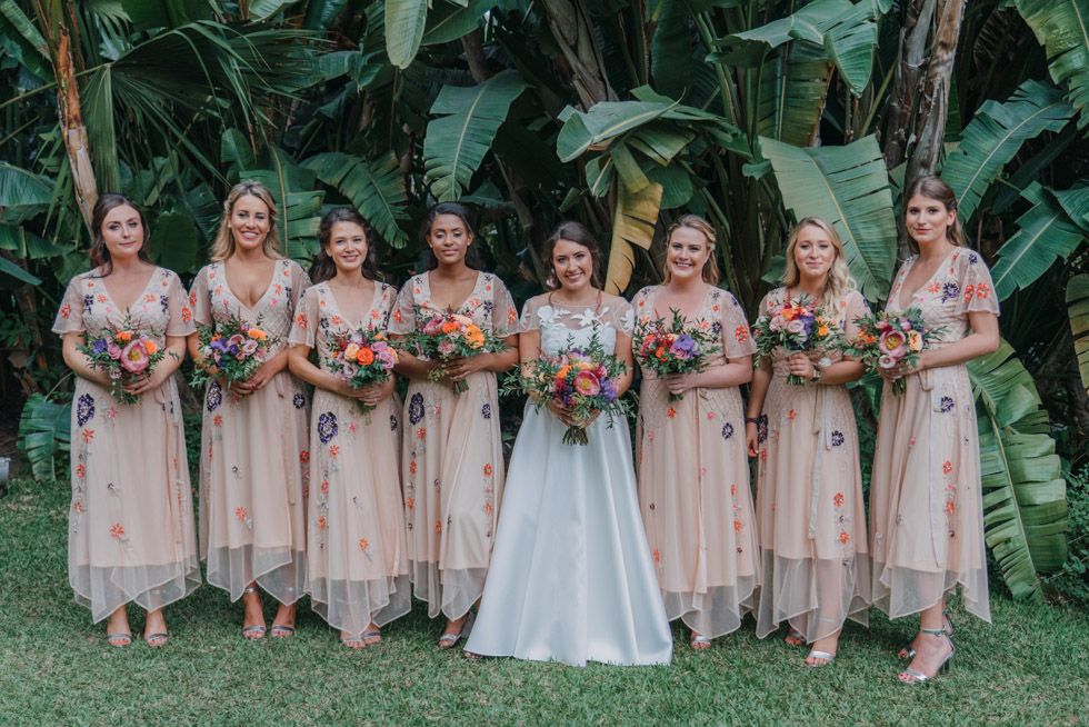 Bridal party in matching pink dresses holding colorful bouquets poses against green tropical backdrop.