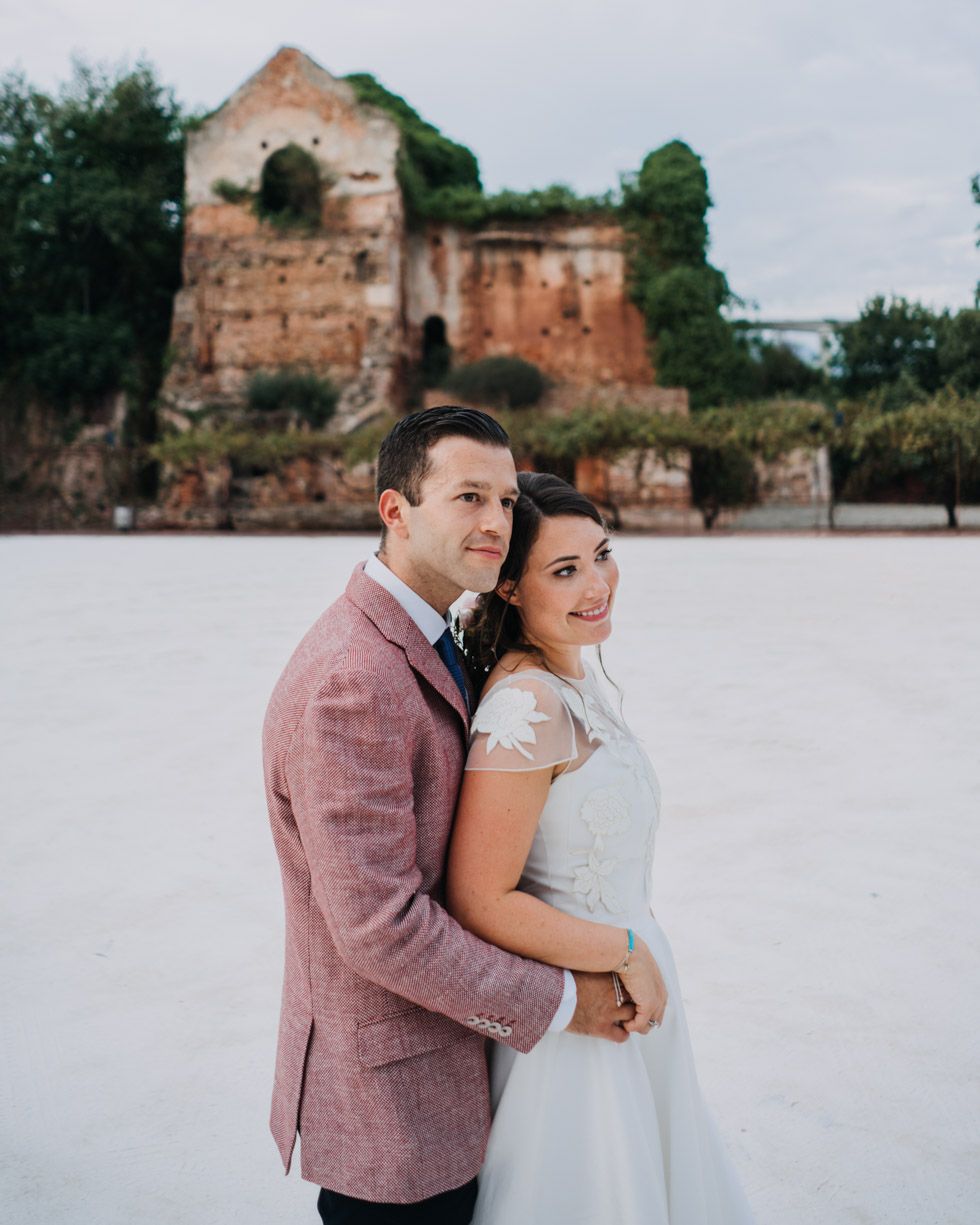 A couple in wedding attire poses together in front of an old stone building with weathered walls.
