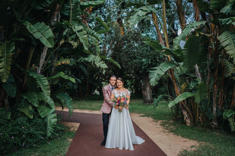 A couple in wedding attire stands together on a brick path surrounded by lush tropical foliage and banana plants.