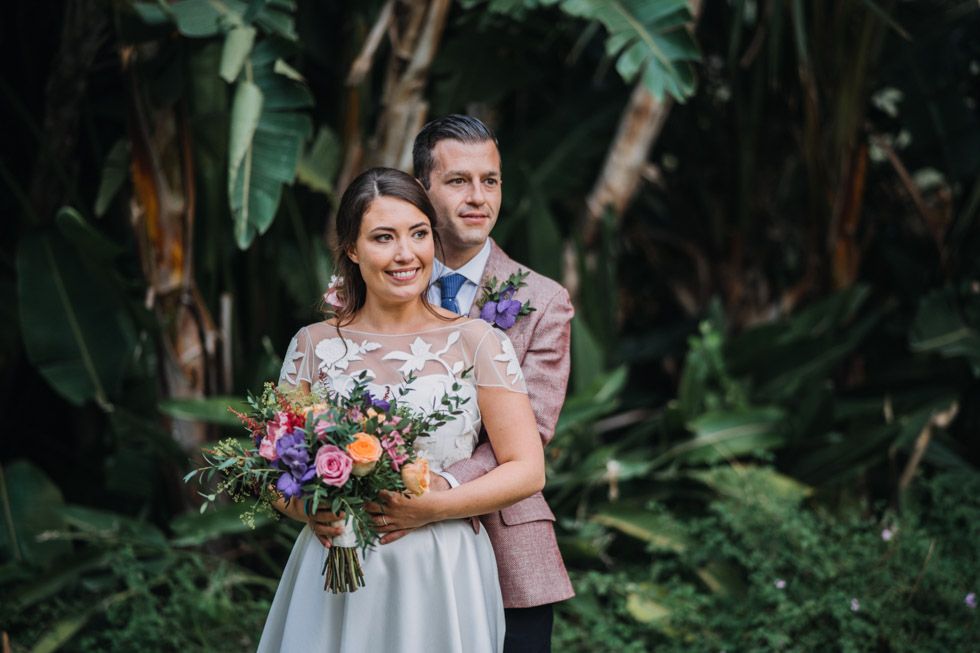 A couple poses together in a lush tropical garden setting with the bride holding a vibrant purple and green bouquet.