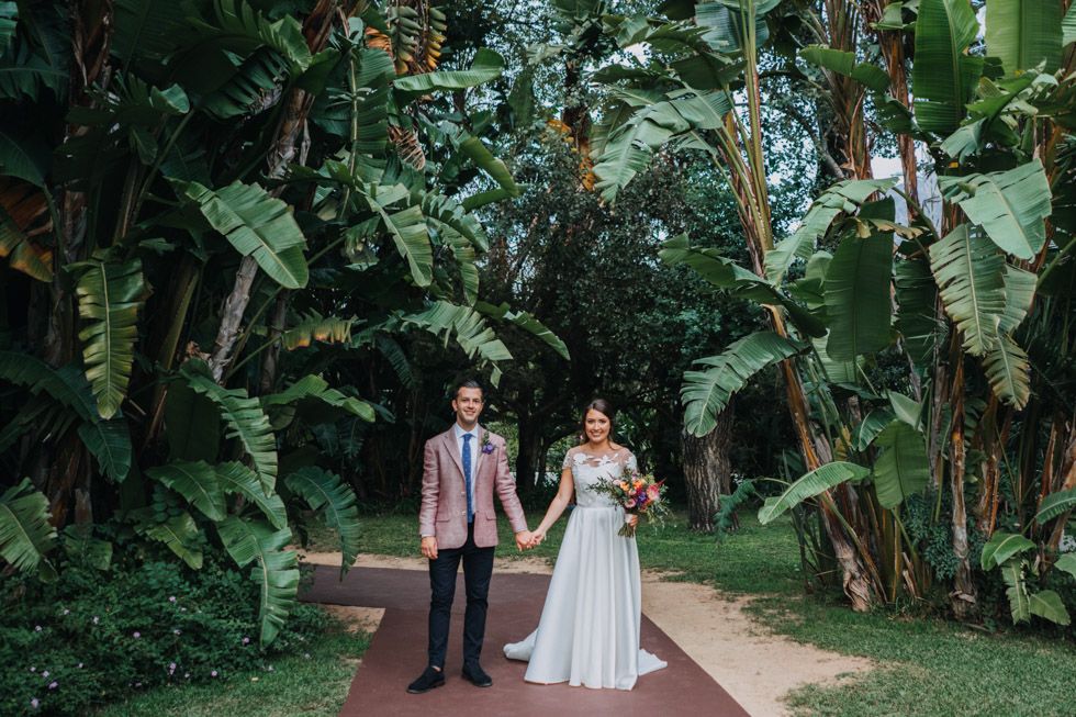 A wedding couple holds hands while walking along a red path surrounded by lush tropical banana trees and greenery.