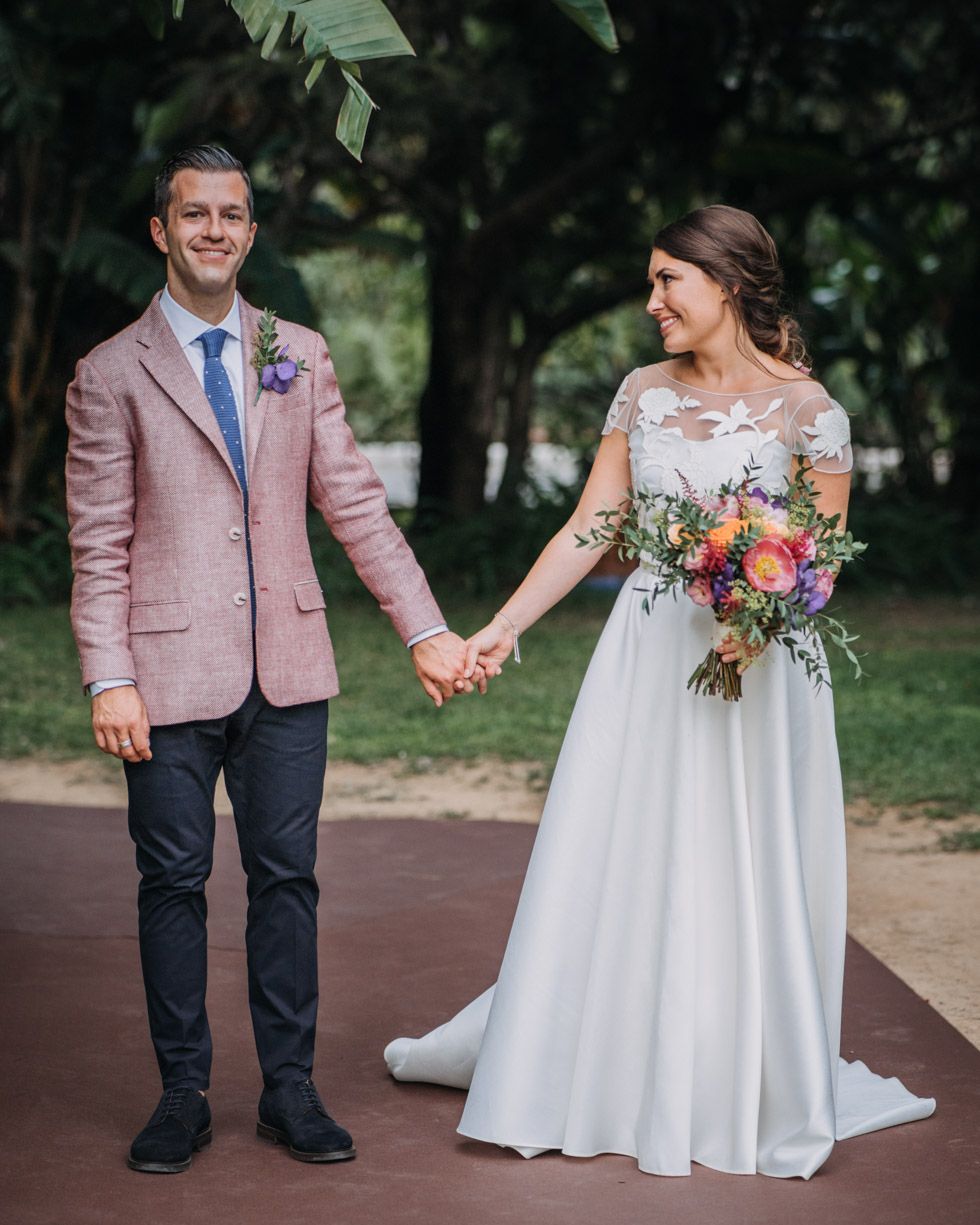 A wedding couple stands together on a path, with bride in white dress and bouquet and groom in pink blazer and navy pants.