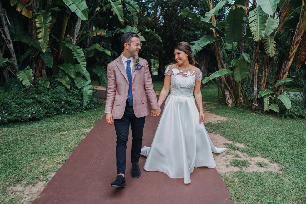 A bride and groom walk hand in hand down a garden path surrounded by lush tropical foliage.
