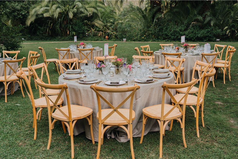 Elegant outdoor wedding reception setup with round tables and wooden crossback chairs on a manicured lawn.