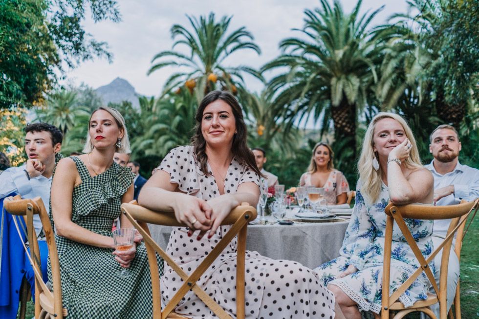 Wedding guests sharing a joyful moment together on wooden chairs with palm trees in the background.
