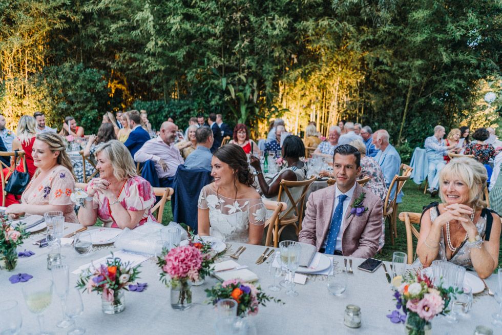 Wedding guests seated at elegant outdoor reception tables with pink floral centerpieces beneath trees at dusk.